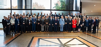 Una foto grupal de aproximadamente sesenta personas, la mayoría hombres en trajes formales, posando en un interior moderno con grandes ventanales
