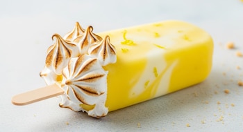 Primer plano de una paleta helada de pay de limón con crema amarilla y blanca veteada, merengue tostado en un extremo y un palo de madera.