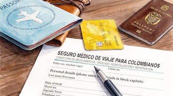 Un formulario de seguro médico de viaje, un bolígrafo, un pasaporte colombiano de color café, una tarjeta de crédito amarilla y un documento de viaje azul.
