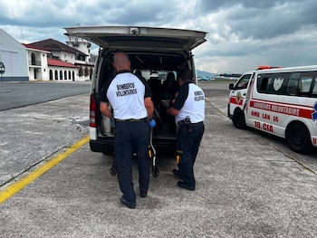 Dos bomberos voluntarios cargan equipo en la parte trasera de una camioneta blanca con el maletero abierto, junto a una ambulancia estacionada en un aeropuerto