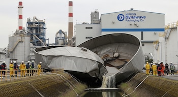 Imagen de una fábrica industrial con un tanque metálico colapsado derramando líquido. Bomberos y personal de seguridad observan. Al fondo, estructuras y logo Nippon Dynawave.