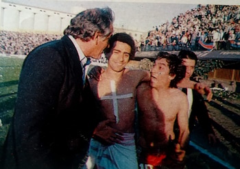 Cuatro hombres en un estadio, uno con traje, tres semidesnudos. Uno lleva una cruz blanca en el pecho. Fondo de gradas con público