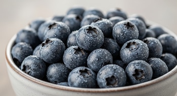 Primer plano de un tazón de cerámica lleno de arándanos frescos y redondos, cubiertos de pequeñas gotas de agua, sobre un fondo claro.