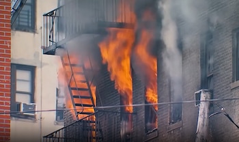 Vista exterior de un edificio de ladrillo con grandes llamas y humo denso que salen de múltiples ventanas y una escalera de incendios
