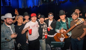 Un grupo de hombres posa en un recinto oscuro, algunos portan sombreros o gorras, vasos rojos y una guitarra