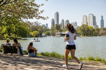 Una mujer corre por un camino junto a un lago mientras varias personas descansan en bancos con árboles y edificios de fondo.