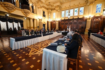 Interior de un salón con arquitectura clásica, columnas, balcones y ventanales con vitrales. Múltiples personas sentadas en mesas largas con micrófonos