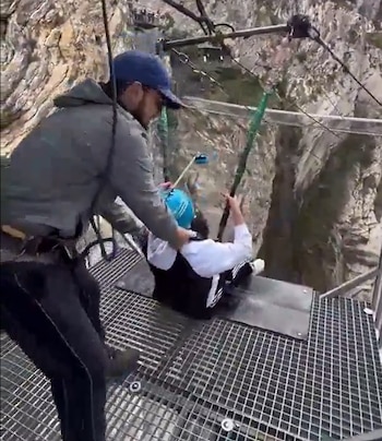 Un hombre con gorra ajusta el arnés de una persona con casco sentada al borde de una plataforma de metal frente a un cañón rocoso