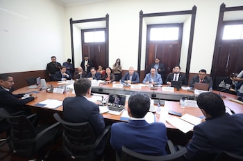 Personas en traje formal sentadas alrededor de una mesa ovalada de madera con computadoras portátiles, micrófonos y documentos