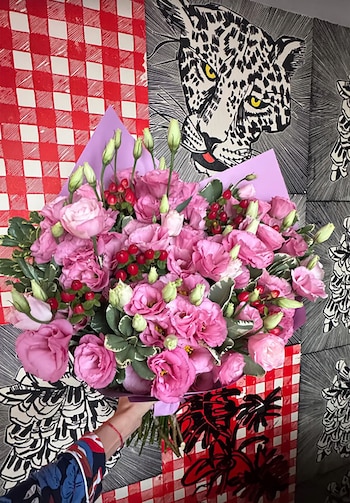 Una mano sostiene un gran ramo de flores rosadas, capullos verdes y bayas rojas, envuelto en papel morado, frente a un fondo estampado con cuadros y un leopardo