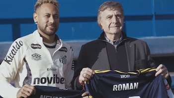 Neymar, con camiseta blanca del Santos FC, sonríe junto a un hombre mayor que sostiene una camiseta azul oscuro con el nombre "ROMAN"