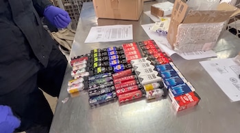 Una persona con guantes morados junto a una mesa metálica con múltiples botellas pequeñas, cajas de cartón abiertas, y documentos