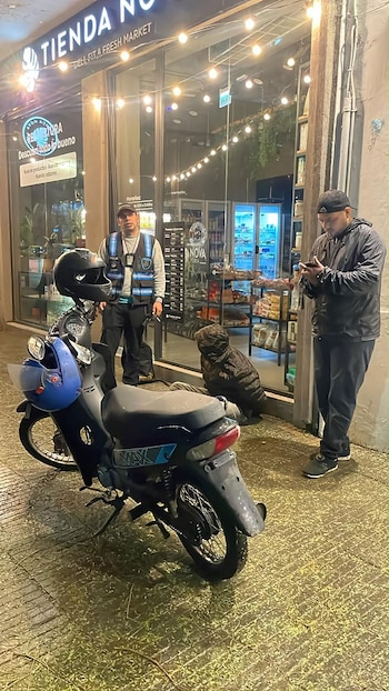Un oficial de seguridad permanece junto a una persona sentada en la vereda y una motocicleta estacionada frente a un comercio de noche