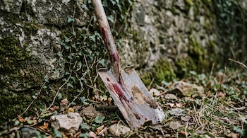 Una pala con manchas rojas y tierra apoyada contra un muro de piedra con vegetación en el suelo.