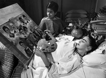 Fotografía en blanco y negro de Frida Kahlo acostada en la cama con una paleta y pinceles frente a un caballete, observada por un niño