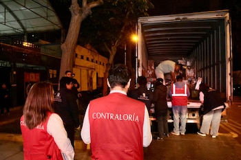 Vista trasera de personas con chalecos de "Contraloría" y "ONPE" supervisando la descarga de cajas de un camión grande por la noche