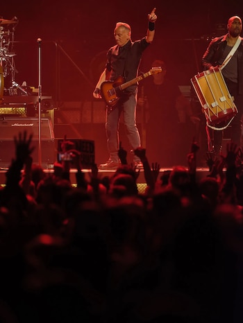 Bruce Springsteen de pie en un escenario oscuro, cantando en un micrófono y tocando una guitarra eléctrica color crema. Luces verdes iluminan la batería detrás de él