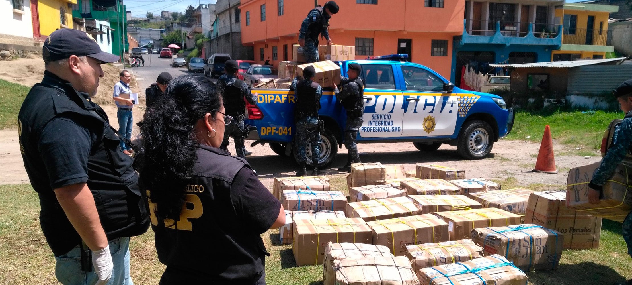 Agentes de policía y personal del Ministerio Público supervisan la descarga de múltiples cajas de cartón de un vehículo policial, dispuestas sobre el pasto en una zona urbana. (Agencia Guatemalteca de Noticias)