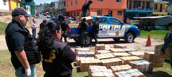 Oficiales de policía, personal del Ministerio Público, un vehículo policial azul, cajas de cartón en el suelo y siendo cargadas, casas y calle