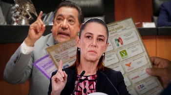 Mujer con cabello recogido, saco negro y blusa colorida, gesticula con el índice hacia arriba ante un micrófono; un hombre difuminado y papeletas electorales al fondo.