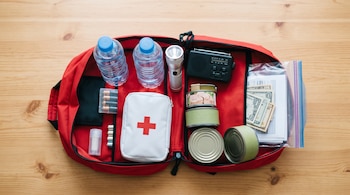 Mochila roja abierta sobre madera con botellas de agua, linterna, radio, botiquín, latas de comida, pilas y dinero en bolsa plástica.