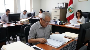 Hombre adulto mayor con gafas revisa documentos en una oficina con otros funcionarios, computadoras, escritorios, archivos y una bandera de Perú.