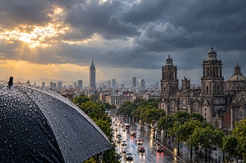 Un paraguas negro mojado en primer plano frente a una avenida con tráfico bajo la lluvia, un templo catedralicio y un rascacielos al fondo