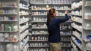 Uma mulher por trás pega um frasco de remédio de uma prateleira. As prateleiras das farmácias estão cheias de centenas de frascos e caixas de remédios