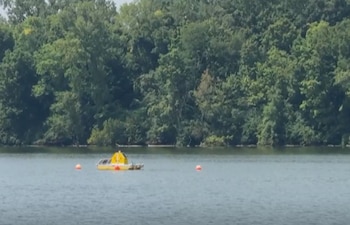 En un río, un dispositivo flotante amarillo con dos boyas anaranjadas a sus lados, y un fondo de densa vegetación con árboles verdes