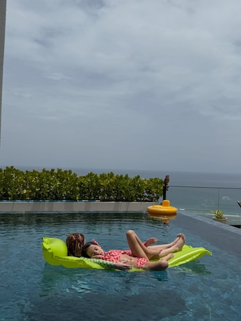 Niña con trenzas y traje de baño rojo a cuadros flota en un colchón inflable verde en una piscina. Fondo con mar, cielo nublado, flotador amarillo y un pájaro