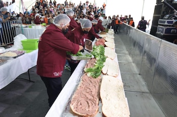 Varias personas con uniformes, cofias y guantes preparan una torta gigante con pan, untable y lechuga, ante una multitud que observa