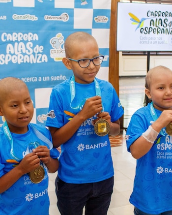 Tres niños con camiseta azul y cabeza rapada sujetan medallas doradas. Un niño con gafas, otro con brazo vendado. Pancartas de evento al fondo