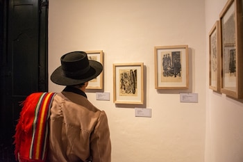 Una persona de espaldas con sombrero negro y poncho a rayas contempla varios cuadros con grabados colgados en una pared blanca