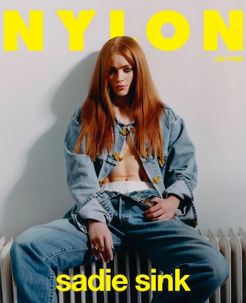 Mujer joven de cabello rojizo, con chaqueta y pantalón de mezclilla, sentada sobre un radiador. Fondo blanco. Texto: NYLON, JULIO 2023, sadie sink