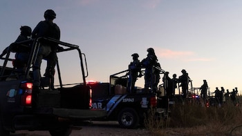 Agentes de seguridad en silueta, con cascos y equipo táctico, de pie en camionetas pickup oscuras con luces de emergencia. Fondo de cielo al anochecer