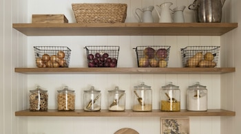 Tres estantes flotantes de madera clara en una pared blanca con tablillas. Contienen cestas de metal con cebollas, patatas y calabazas, y frascos de vidrio con galletas, cereales y azúcar.