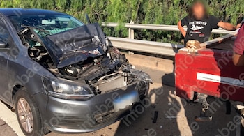 Un automóvil gris con la parte frontal destruida y el parabrisas roto junto a un camión rojo en una carretera.