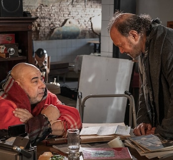 Dos hombres, uno sentado con una manta roja y otro de pie inclinado sobre una mesa con papeles, libros y un vaso