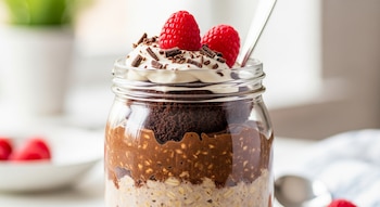 Un frasco de vidrio transparente contiene avena trasnochada de pastel de chocolate, cubierta con crema batida, frambuesas frescas y virutas de chocolate.