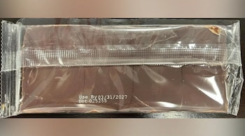 Primer plano de una barra de chocolate de color marrón oscuro dentro de un empaque de plástico transparente. Se lee "Use By 03/31/2027" y "Lot 025255"