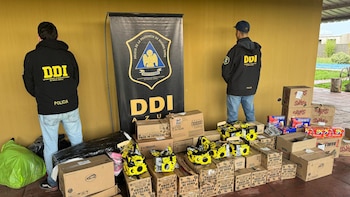 Dos oficiales de la DDI de espaldas frente a un cartel de la Policía de Buenos Aires, junto a pilas de cajas de mercadería recuperada y una bolsa verde