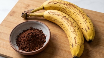 Dos bananas medianas muy maduras con manchas oscuras y un tazón con cacao amargo en polvo sobre una tabla de madera clara.