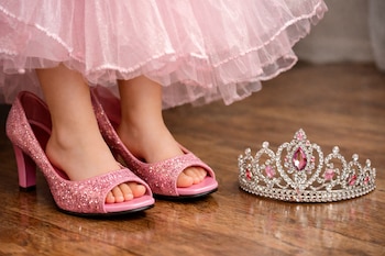 Pies de niña con vestido rosado y tacones brillantes de adultos que le quedan grandes. Una corona plateada con joyas rosadas y blancas está en el suelo de madera.