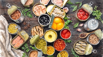 Latas de conserva abiertas con maíz, frijoles, pescado, aceitunas y frutas junto a ajos y hierbas sobre una mesa de madera.