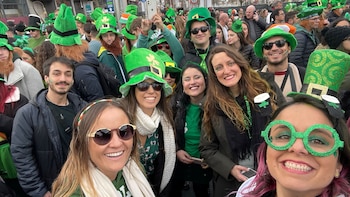 Varias personas sonrientes en una multitud llevan sombreros verdes de copa, gafas con forma de trébol y ropa verde