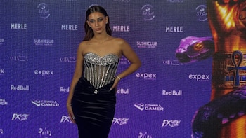Una mujer con cabello oscuro y un top corsé plateado brillante y falda larga negra posa frente a un fondo púrpura con logos de marcas y una figura egipcia