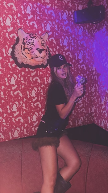 Mujer con gorra negra, chupetín en la boca y un abanico morado brillante, sentada en un sillón rojo. Pared con papel tapiz estampado y cabeza de tigre