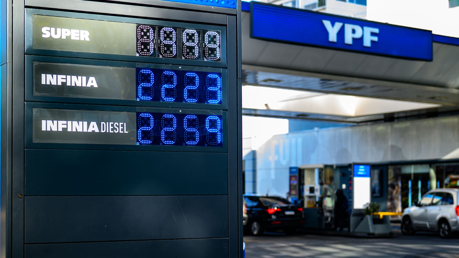 Un cartel de precios de combustible en una estación de servicio YPF muestra los valores actualizados de la nafta Super, Infinia e Infinia Diesel, con la estación y vehículos de fondo. (Adrian Escandar)