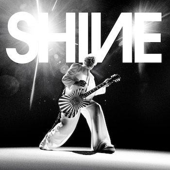 Imagen en blanco y negro de Fito Páez, un hombre con traje claro y gafas de sol, tocando una guitarra con patrón de rayos. La palabra "SHINE" aparece en mayúsculas detrás de él