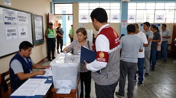 Una mujer deposita su voto en una urna transparente junto a un fiscal, un miembro de mesa y una fila de votantes en una escuela.